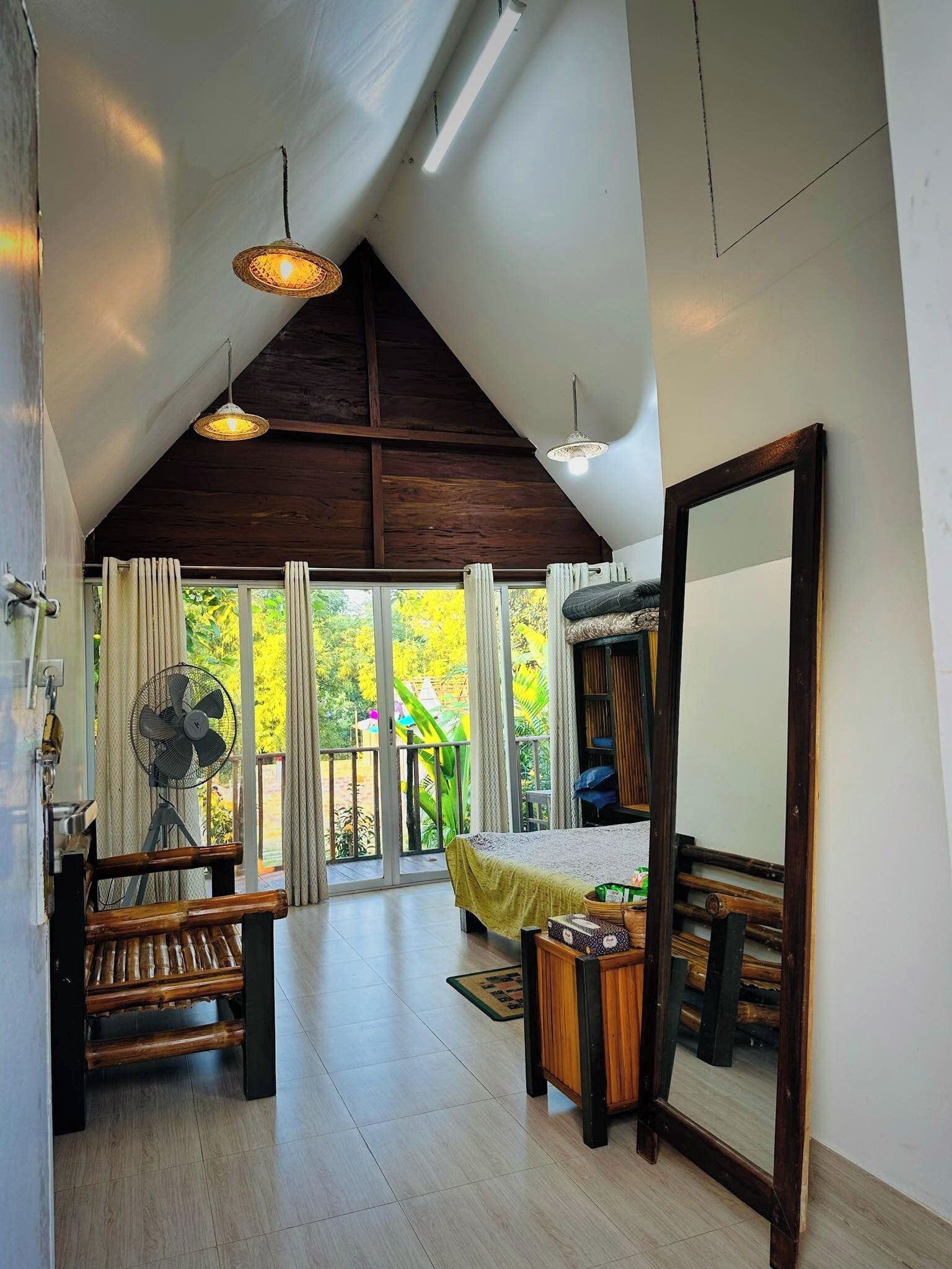 A spacious room with a high ceiling, wooden beams, a large mirror, a fan, a bench, and a view of greenery outside.
