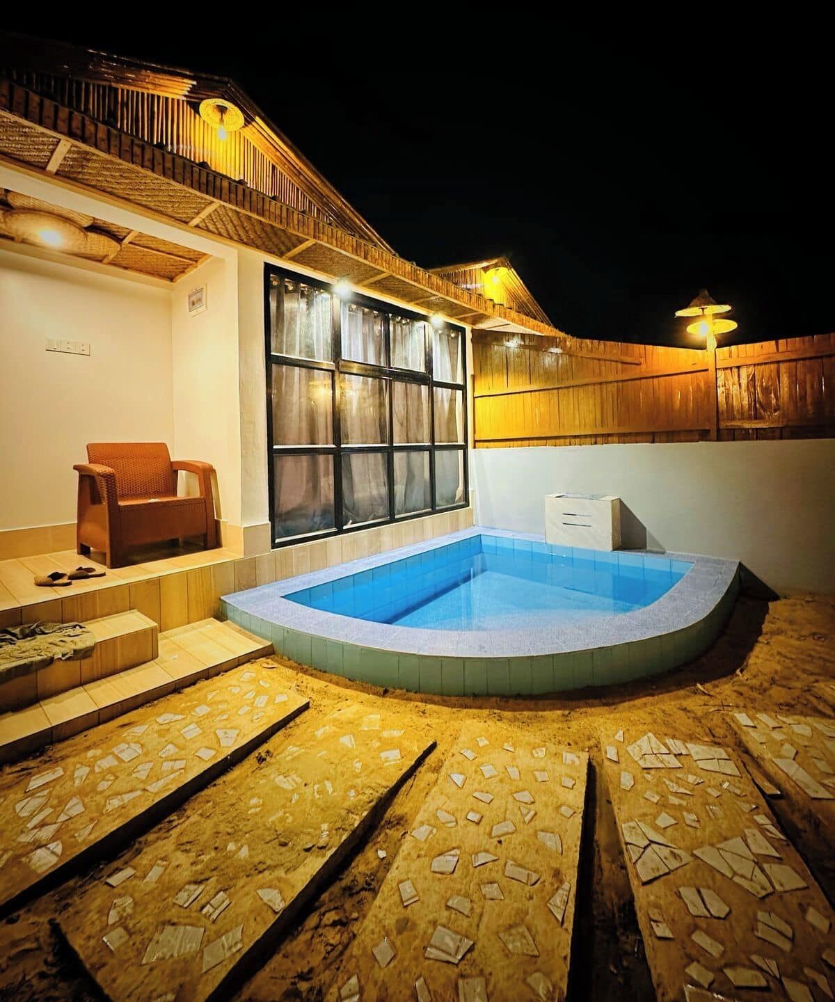 A swimming pool with a wooden deck and steps leading to a covered area with a chair and a small table.