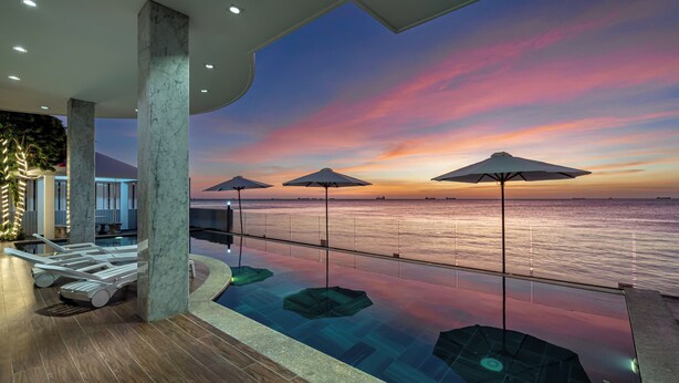 Une piscine intérieure moderne dotée d’un plancher en verre, de grandes colonnes et de parasols, offrant une vue imprenable sur le coucher du soleil.