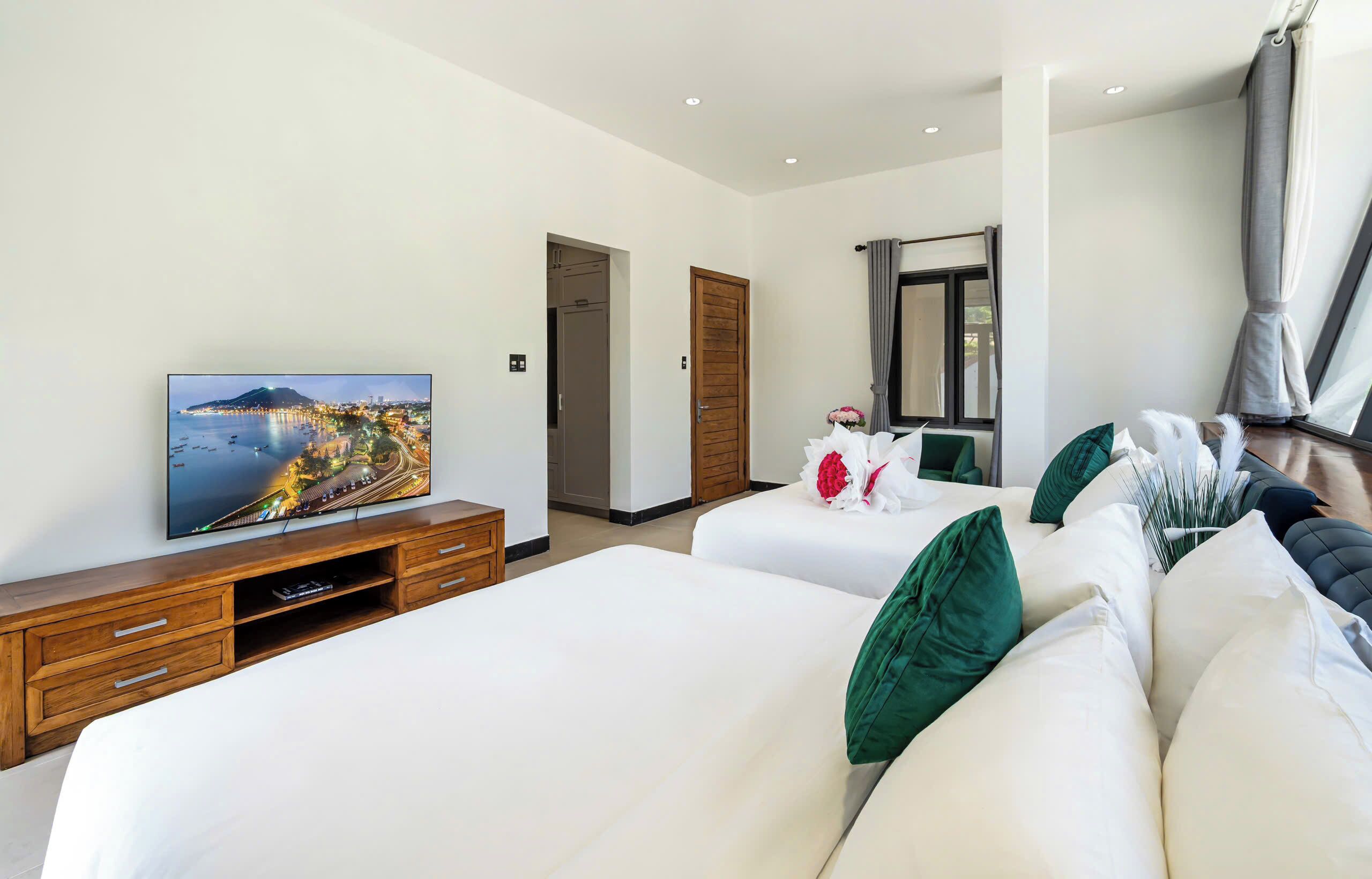 A hotel room with a bed, a TV, a wooden dresser, and a large window with a view of a cityscape.