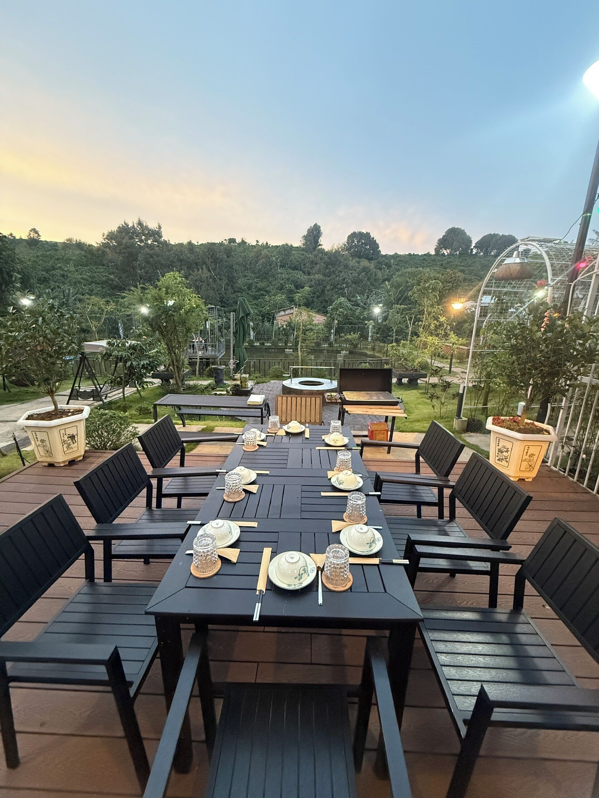 A dining area with a long table set for a meal, surrounded by chairs, with a view of a lush garden and a hill in the background.