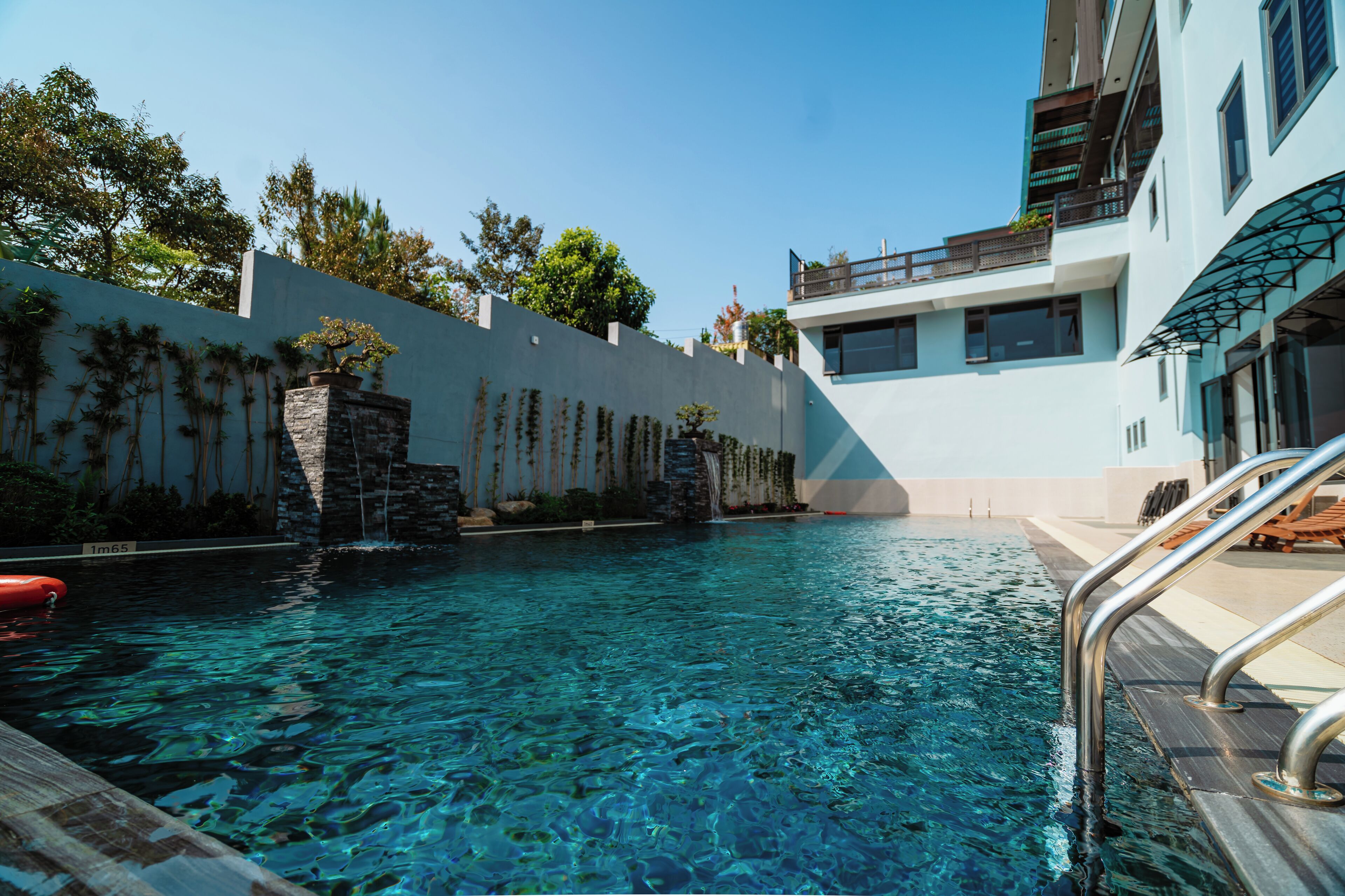 Piscina cristalina con pared de piedra y cascada, rodeada por un edificio blanco con balcones.