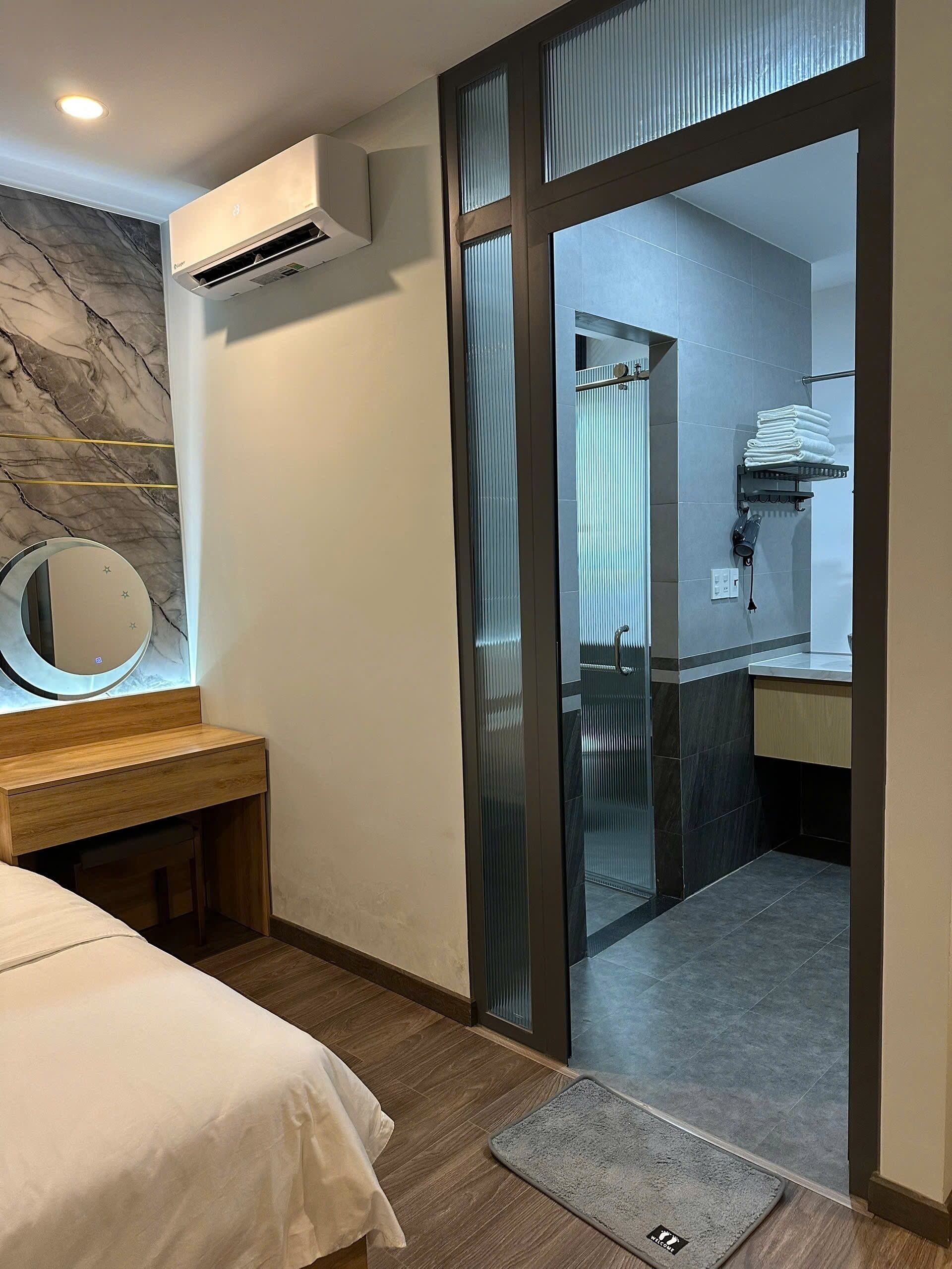 Habitación de hotel con cama, mesita de noche de madera, espejo, equipo de aire acondicionado, baño con ducha y un perchero para toallas.