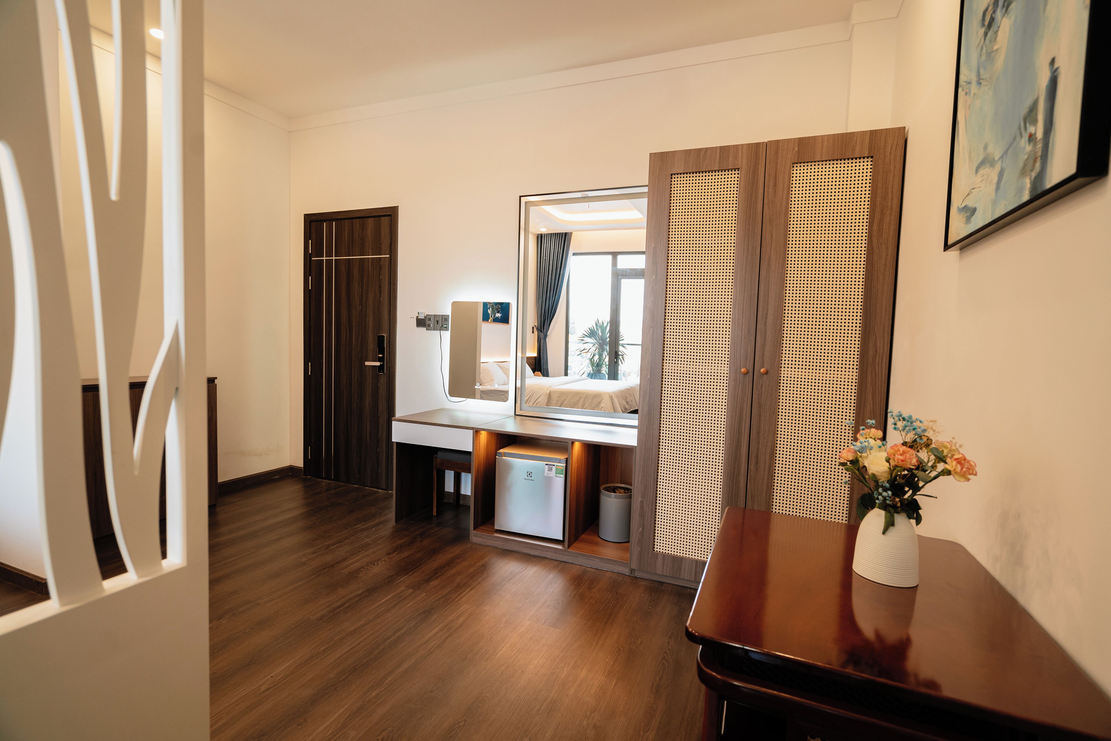 Una habitación de hotel moderna con suelo de madera, un refrigerador pequeño, un espejo, un mueble de madera y un florero con flores sobre una mesa.