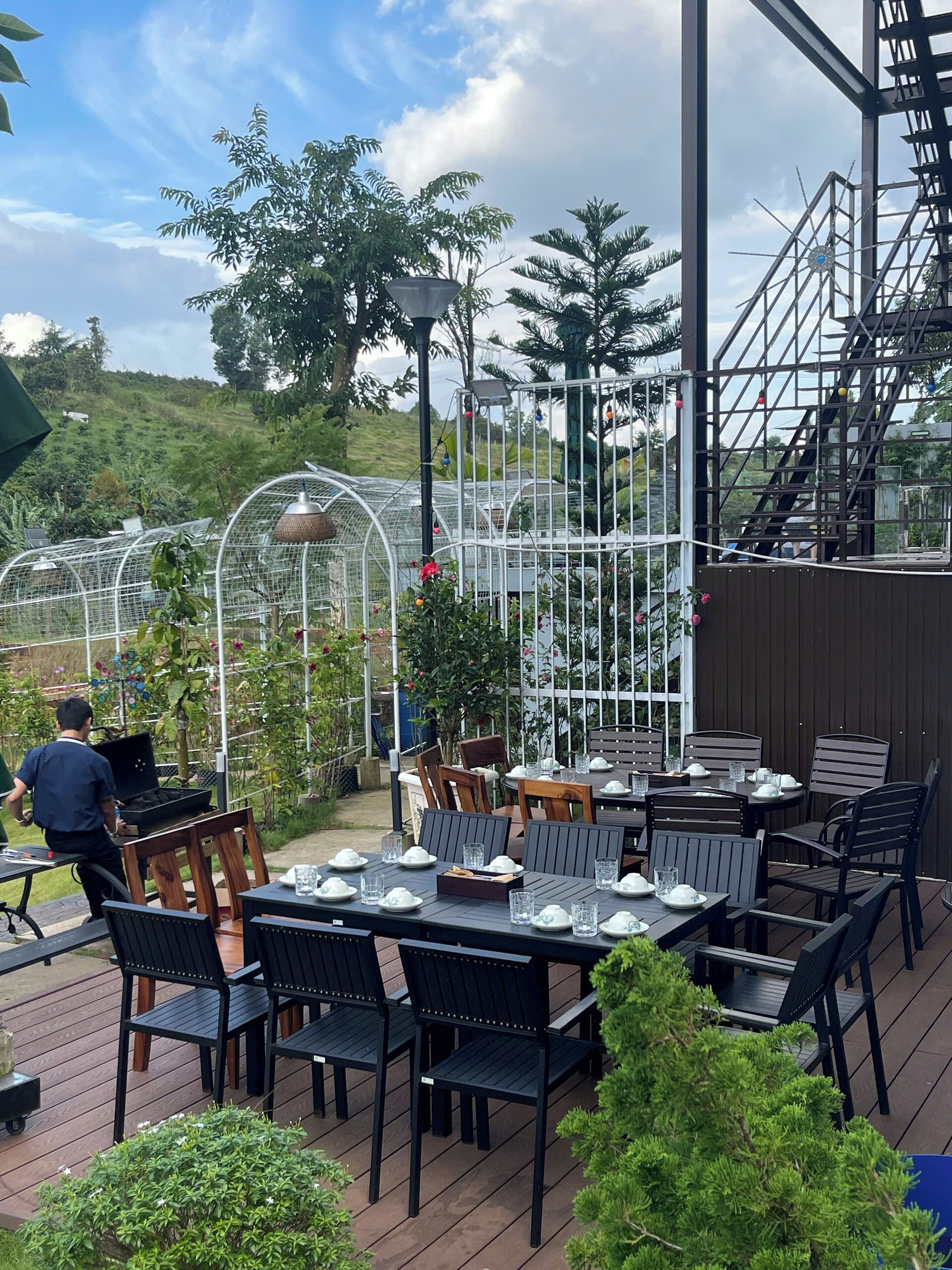 An outdoor patio with tables and chairs, surrounded by greenery and a greenhouse structure.
