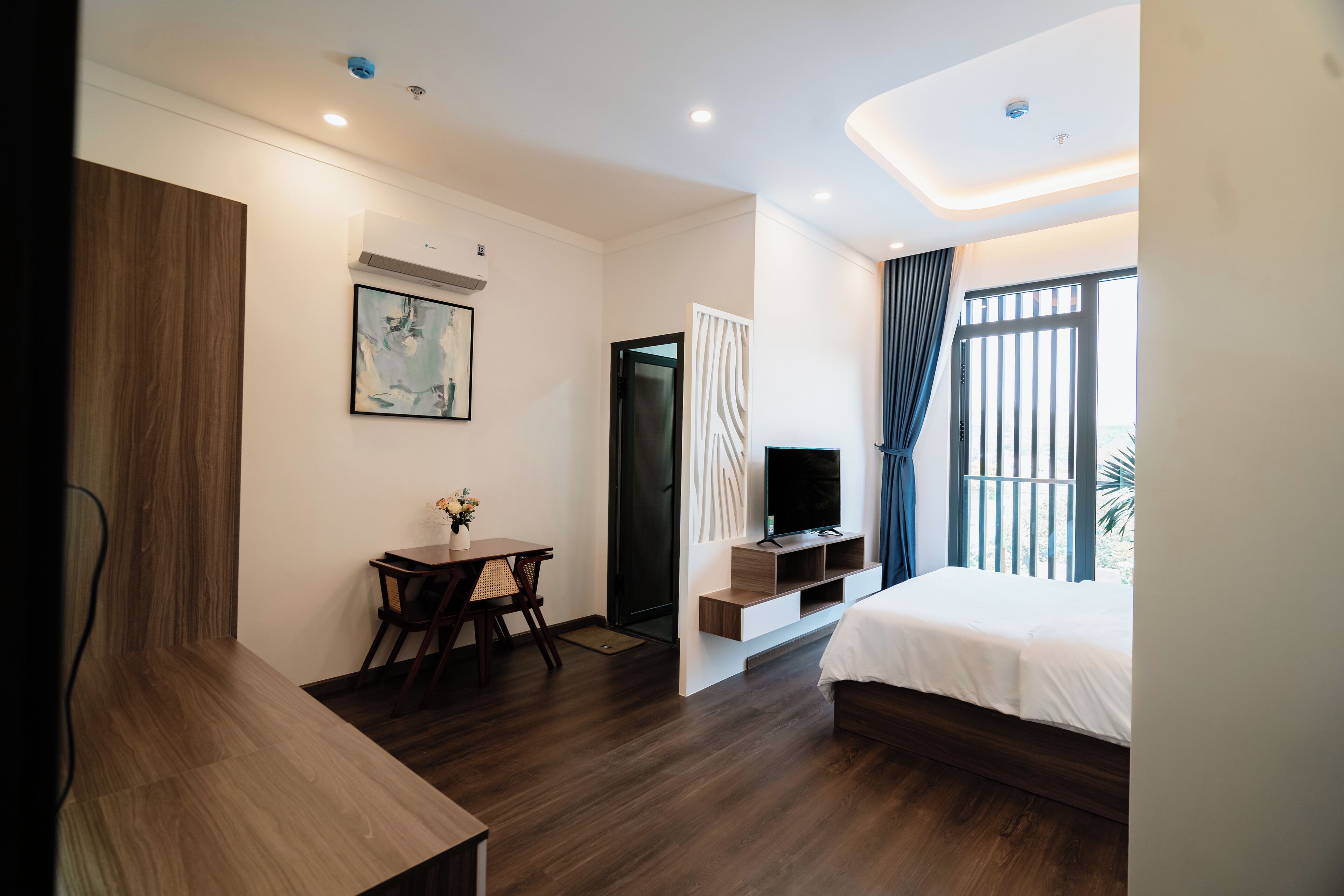 A modern hotel room with a wooden floor, a bed, a desk with a chair, a TV, and a window with curtains.