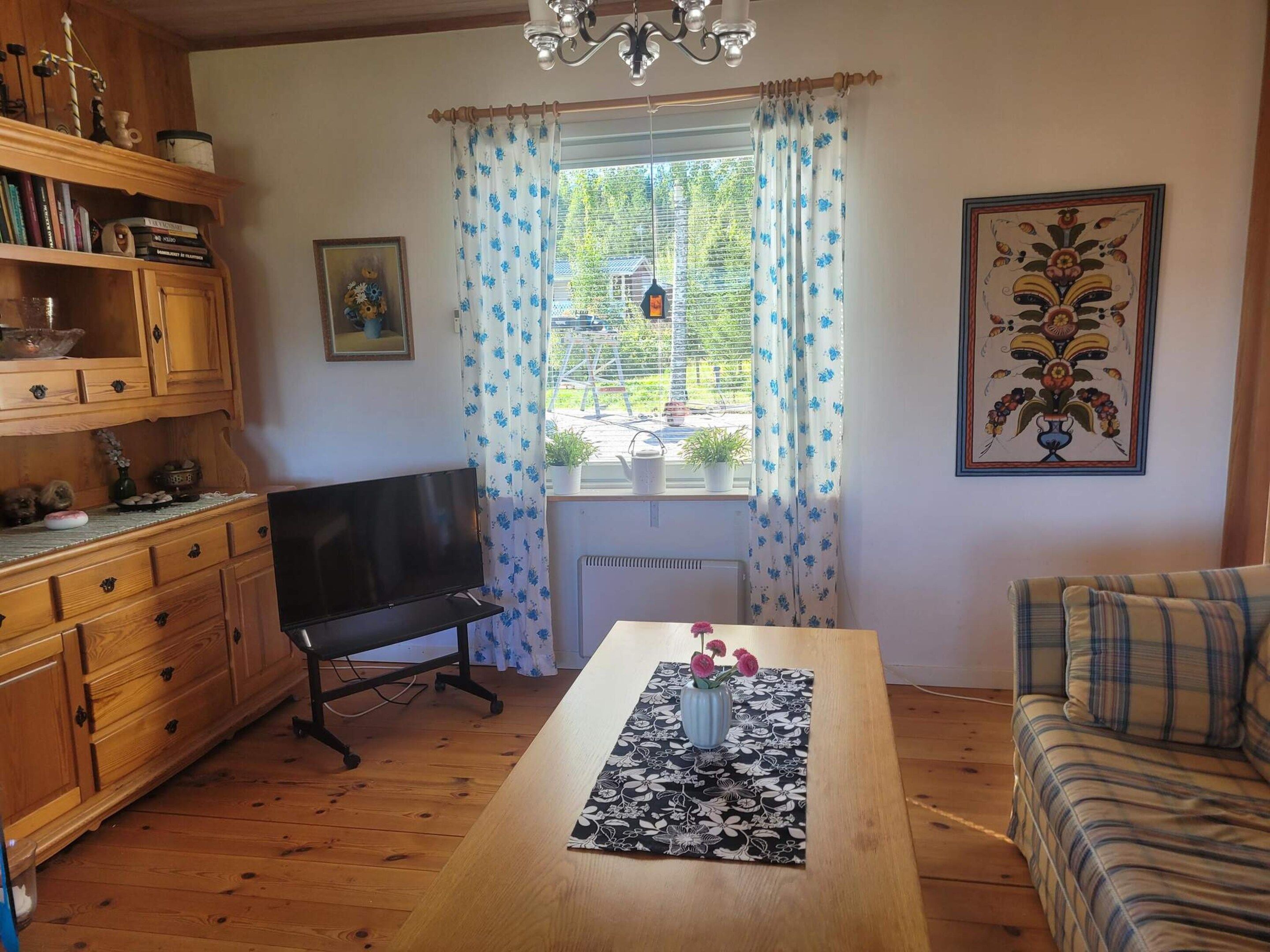A cozy living room with a wooden table, a sofa, a TV, and a window with curtains.