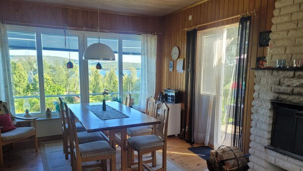 A cozy dining area with a wooden table and chairs, a fireplace, and large windows offering a view of greenery. - 7 Person Holiday Home in Kil (Kil)