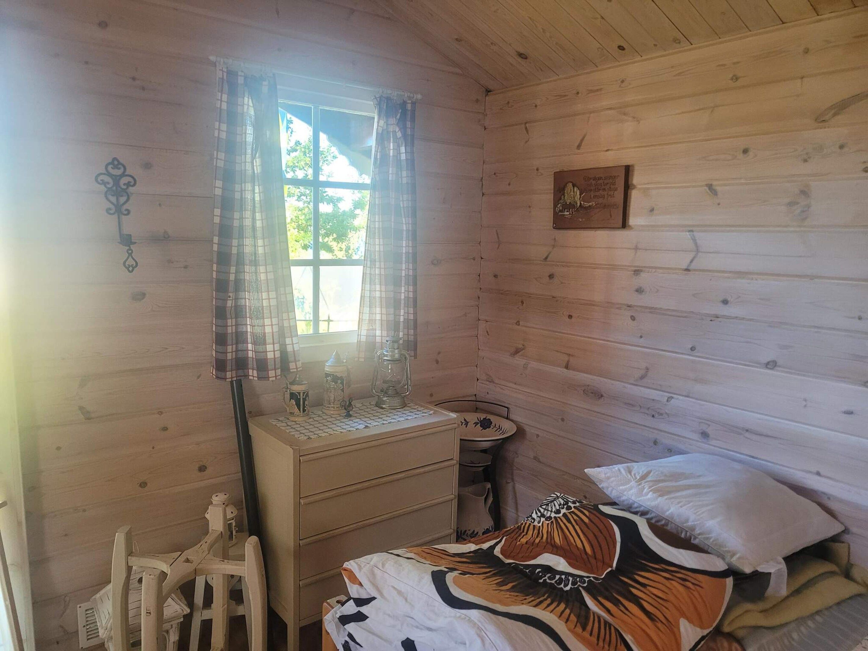 A wooden cabin room with a bed, a small table, a chair, and a window with curtains.