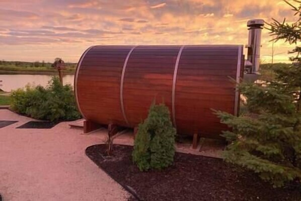 A large wooden barrel-shaped structure with a metal chimney, surrounded by a landscaped garden with small shrubs and a paved walkway. - Lake Front 4 Bedroom Home with Hot Tub & Sauna (Gimli)