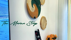 A room with a white wall, a mounted television displaying a beach scene, and a decorative wall piece with the text "The Marina Stays". - The Marina Stays Private Hotel & Spa (Entebbe)