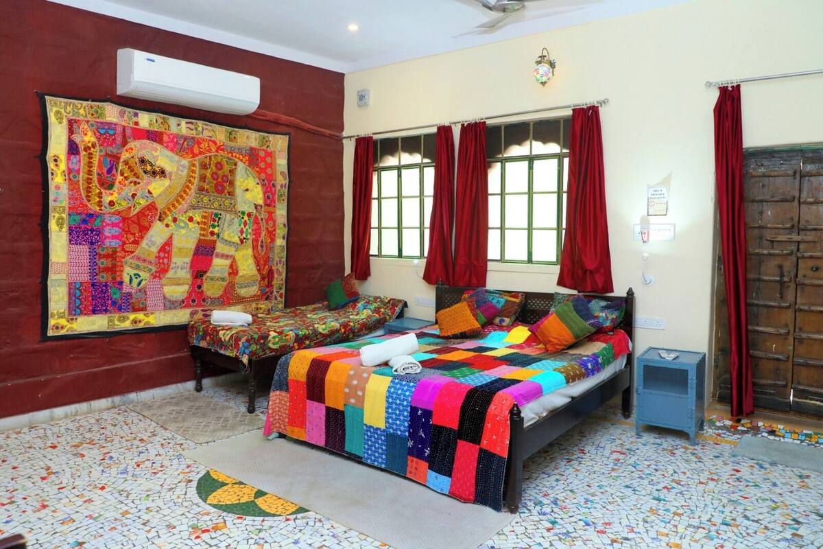 A room with two beds, a large colorful tapestry, a patterned floor, and a traditional wooden door.