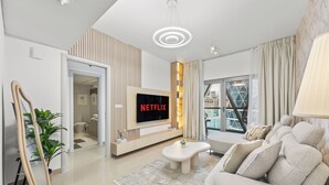Un salon moderne avec un téléviseur à écran plat fixé sur un panneau en bois, un canapé, une table basse et une grande fenêtre donnant sur la ville.