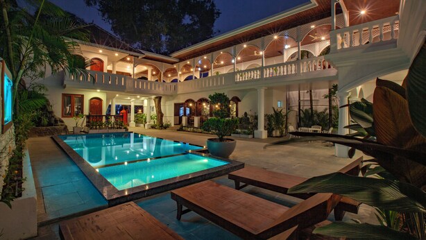 A pool area with wooden loungers, a large pool, and a multi-level building with balconies. - Villa Elixir Fort Kochi (Kochi)