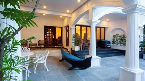 A covered outdoor area with stone flooring, white columns, and seating arrangements. - Villa Elixir Fort Kochi (Kochi)