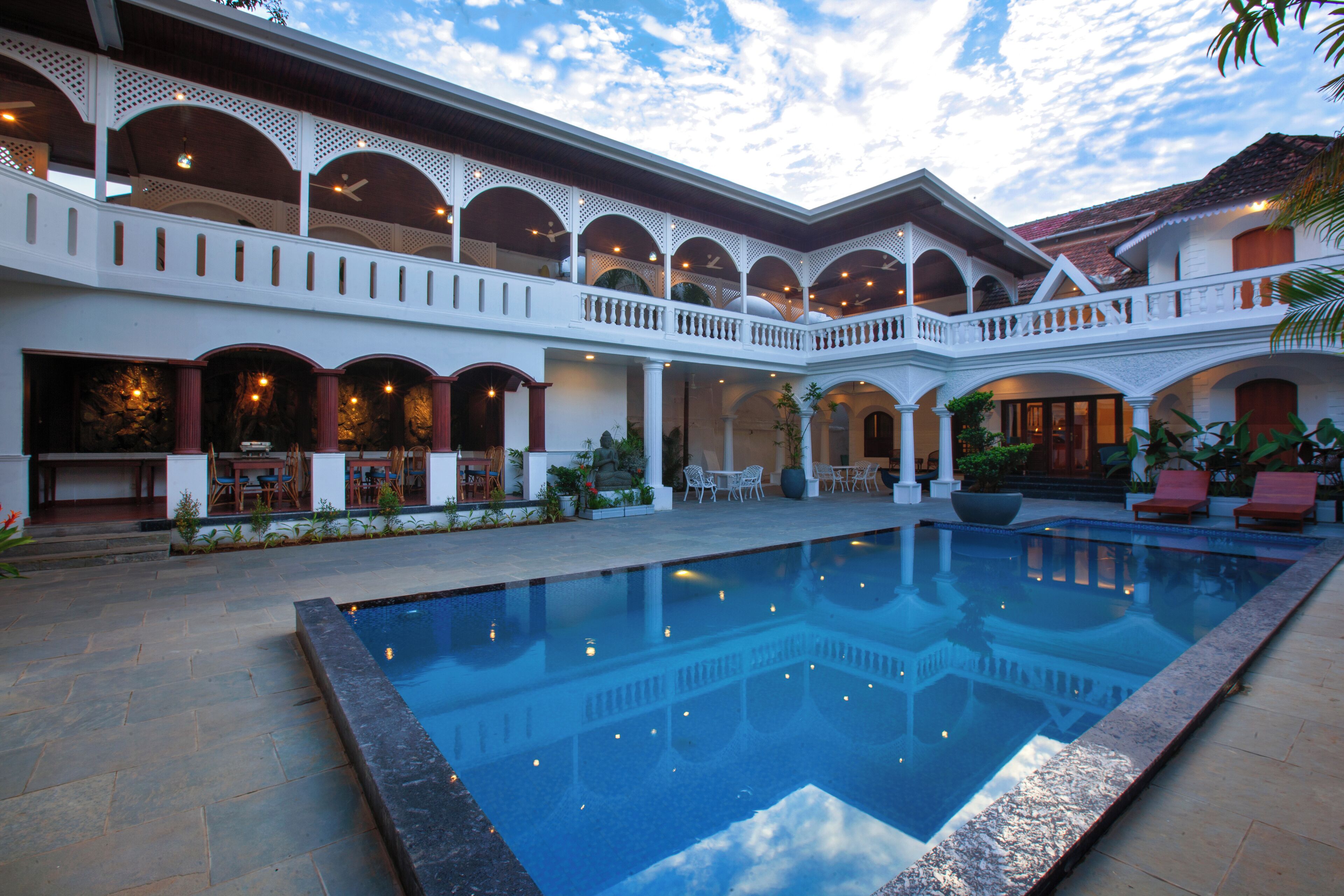 A pool area with a clear blue pool, white columns, and a covered seating area with chairs and tables.