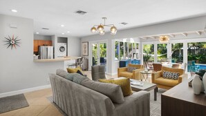 A spacious living area with a grey sofa, yellow armchairs, and a wooden coffee table. A kitchen with stainless steel appliances is visible in the background. - Mid-Century Oasis | Resort Backyard | Near Beach (Fort Lauderdale)