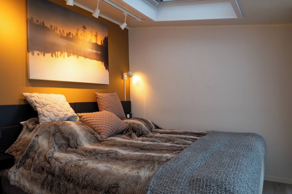 A bedroom with a large bed, a skylight, a painting on the wall, and a bedside lamp.