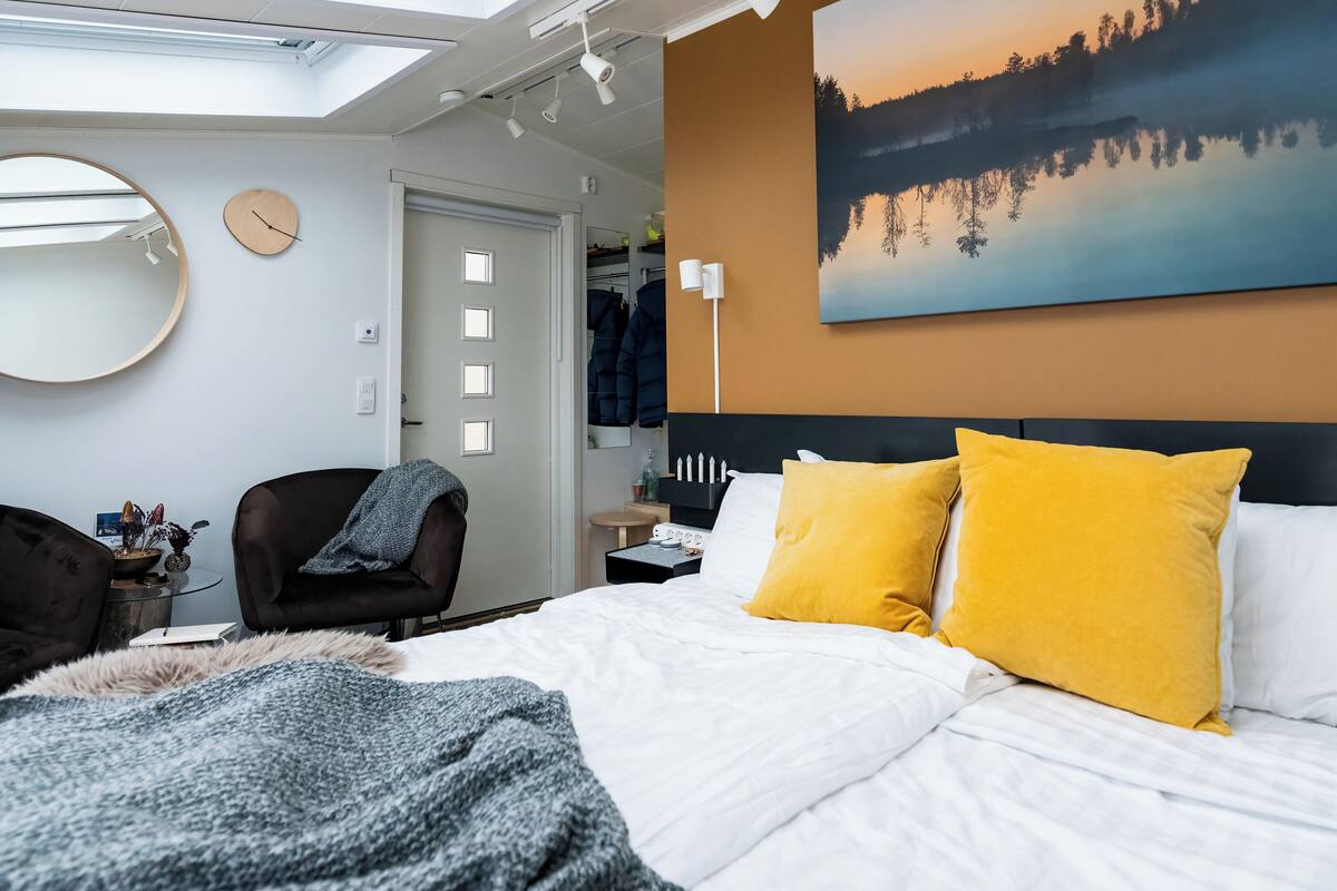 A modern bedroom with a skylight, a large bed with yellow pillows, a round mirror, and a painting of a landscape.