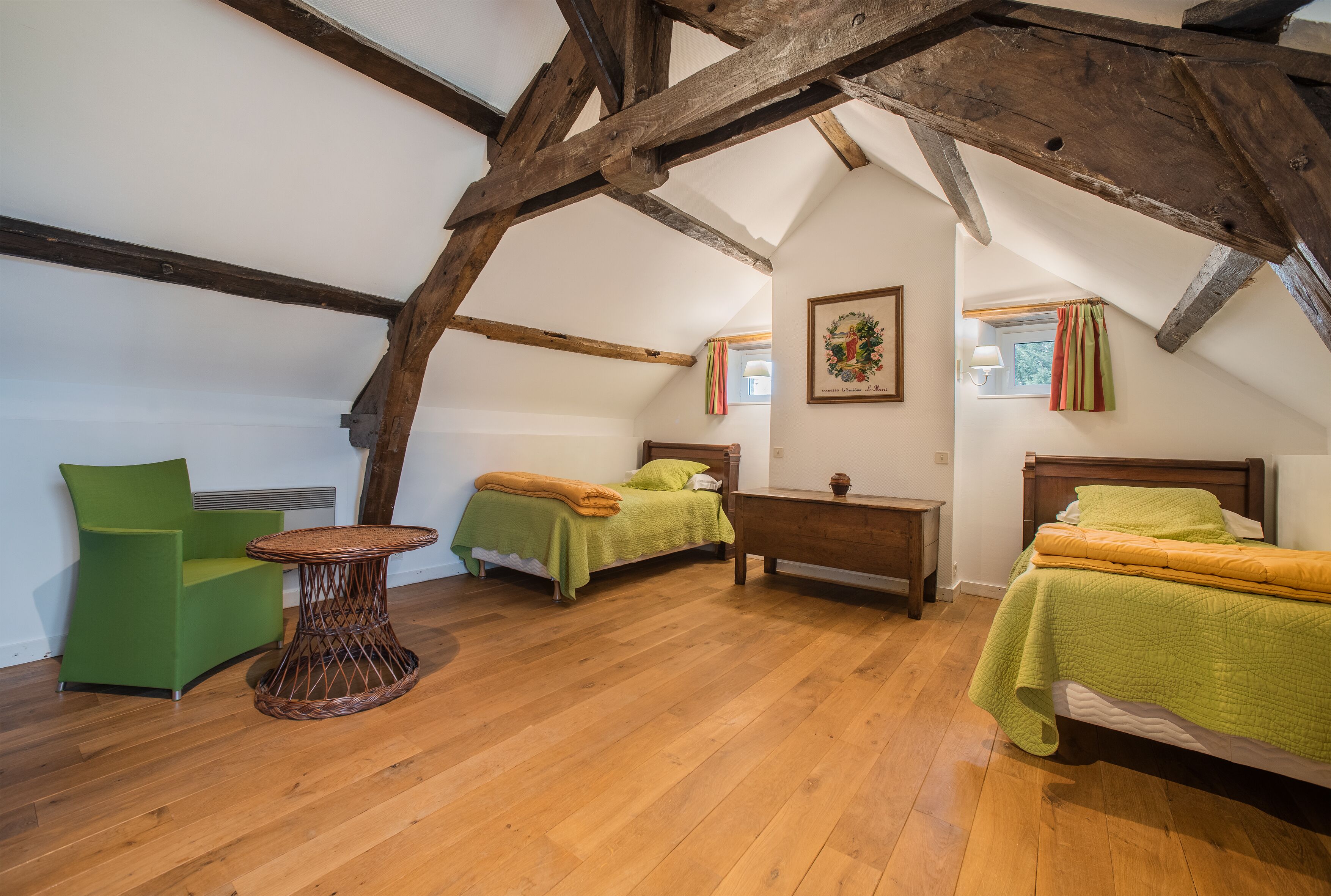A cozy attic bedroom with exposed wooden beams, two single beds with green bedding, a green armchair, and a small round table.
