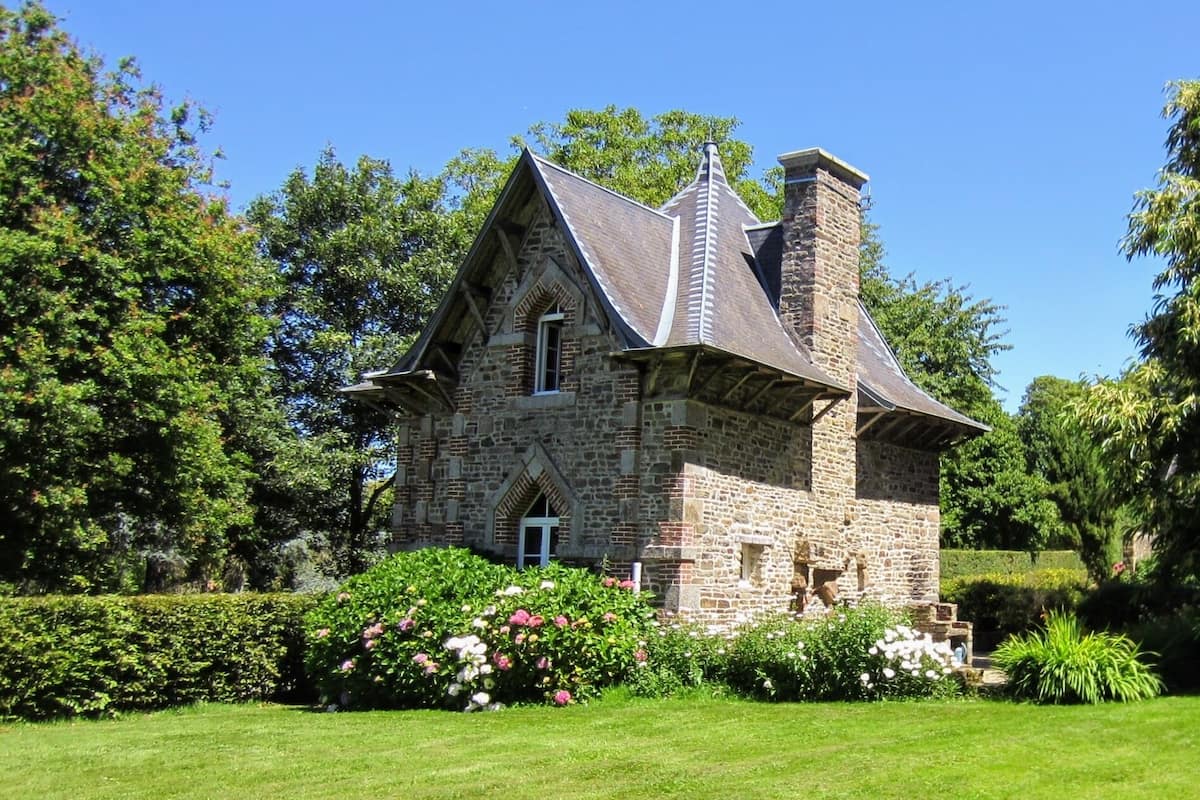 A stone house with a steeply pitched roof and a chimney, surrounded by lush greenery and well-maintained gardens.