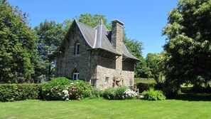 A stone building with a steeply pitched roof and a chimney, surrounded by lush greenery and flowering bushes. - Holiday Home Château de Boucéel Mont Saint Michel (Saint-James)