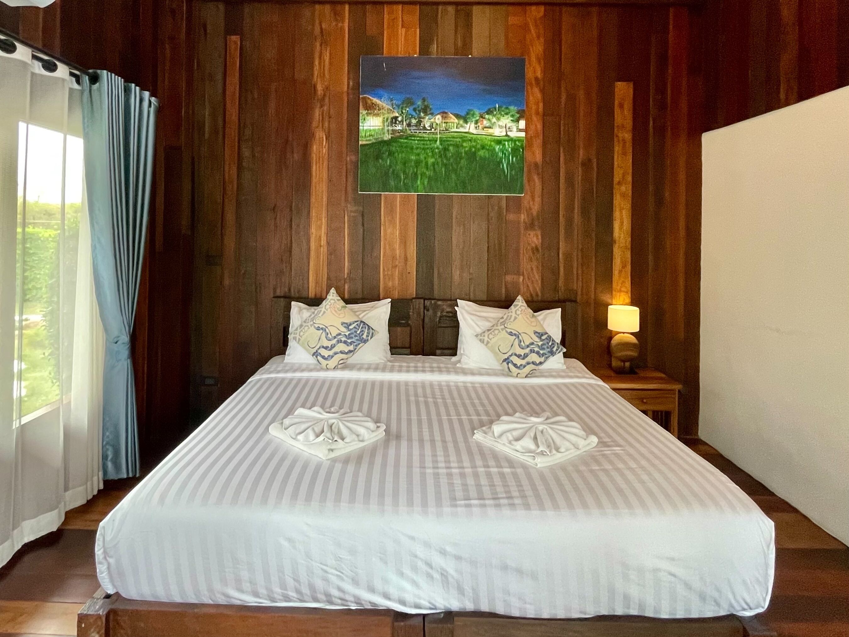 A hotel room with a wooden wall, a bed with white bedding, two pillows with blue patterns, a nightstand with a lamp, and a painting of a landscape.