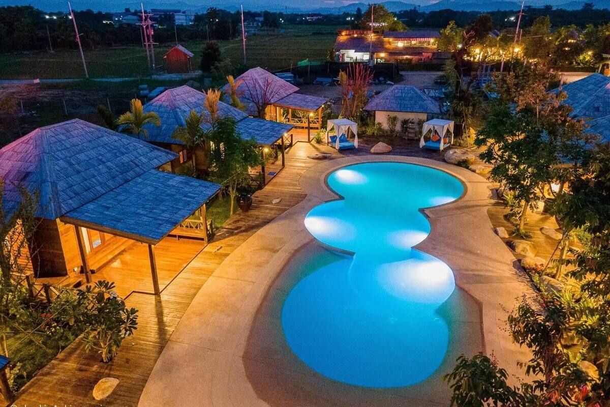 A resort with a large swimming pool, wooden huts, and a well-lit landscape at dusk.