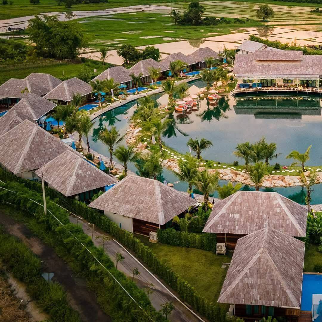 An aerial view of a resort with multiple thatched-roof bungalows arranged around a central swimming pool, surrounded by lush greenery and palm trees.