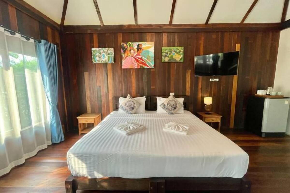 A bedroom with a wooden ceiling, a bed with white linens, two bedside tables, a television, and a small refrigerator.