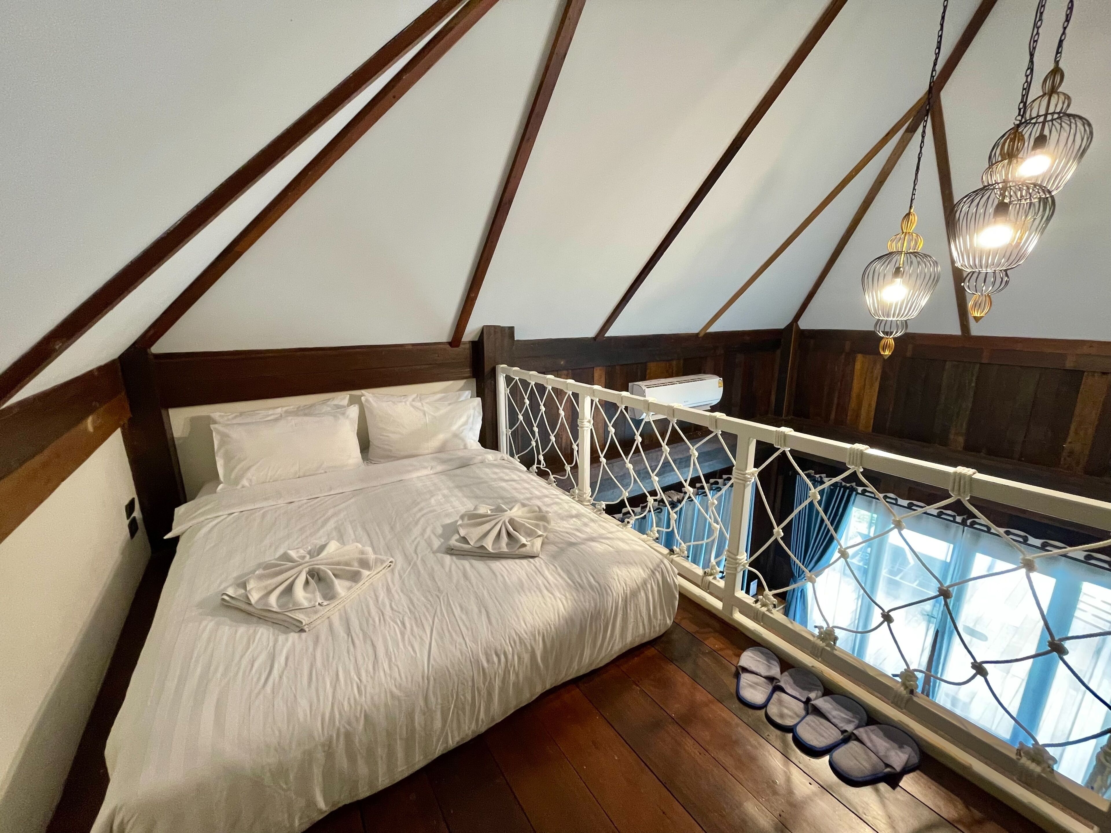 A bedroom with a wooden ceiling, a bed with white linens, a hanging light fixture, and a view through a window with curtains.