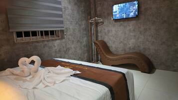 A hotel room with a bed, a brown leather chair, a television mounted on the wall, and a window with blinds.