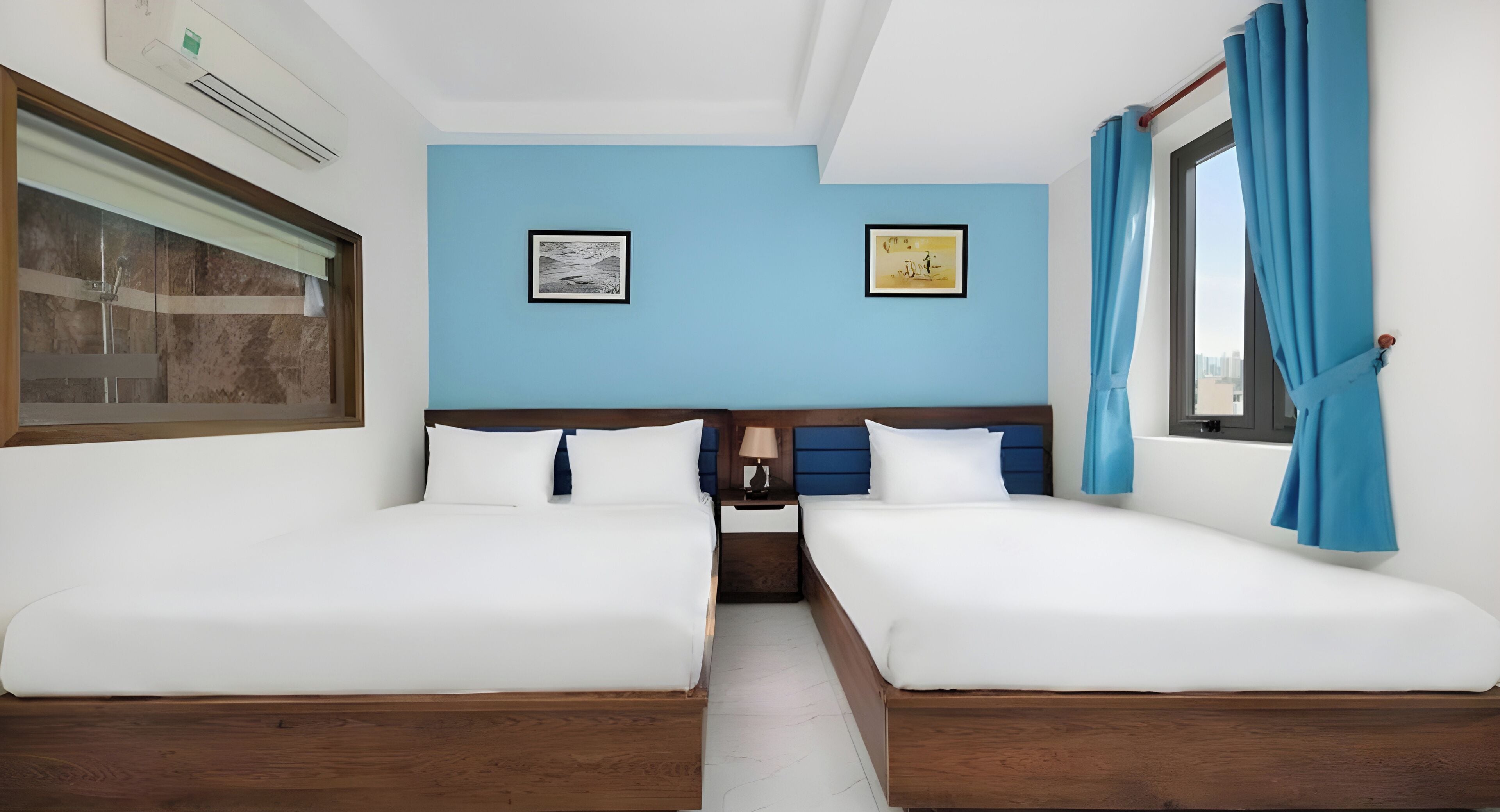 A hotel room with two beds, a window with blue curtains, and two framed pictures on the wall.