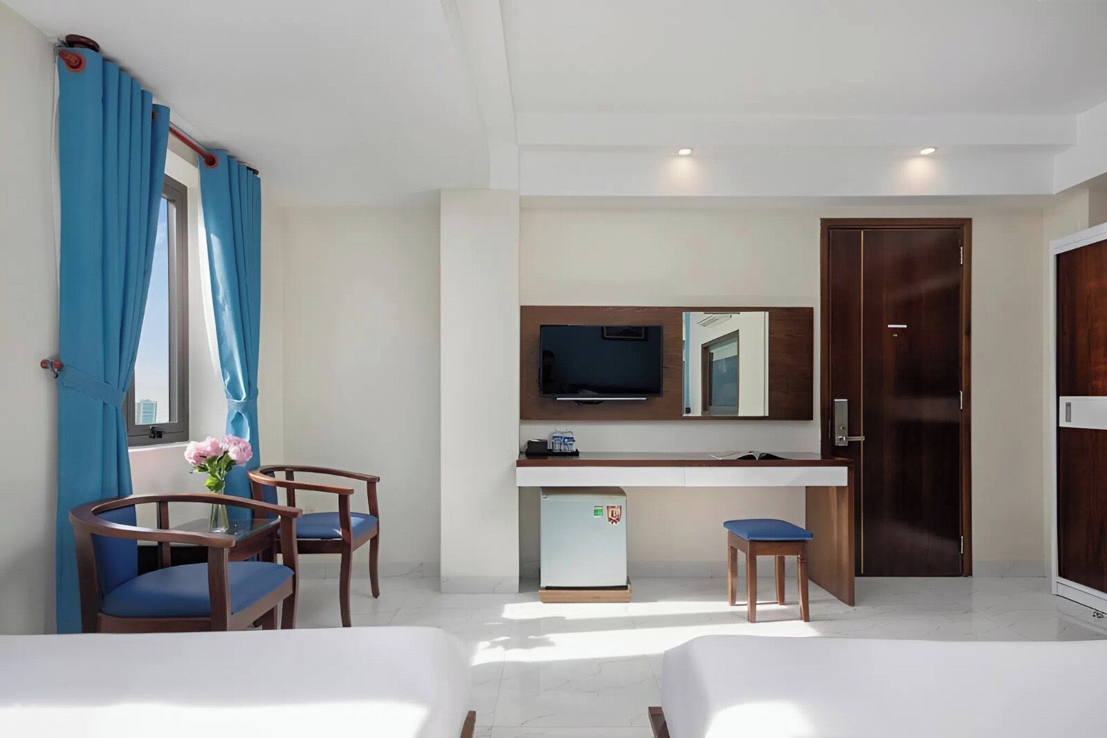 A hotel room with a bed, a desk with a TV, a small fridge, and a window with curtains.