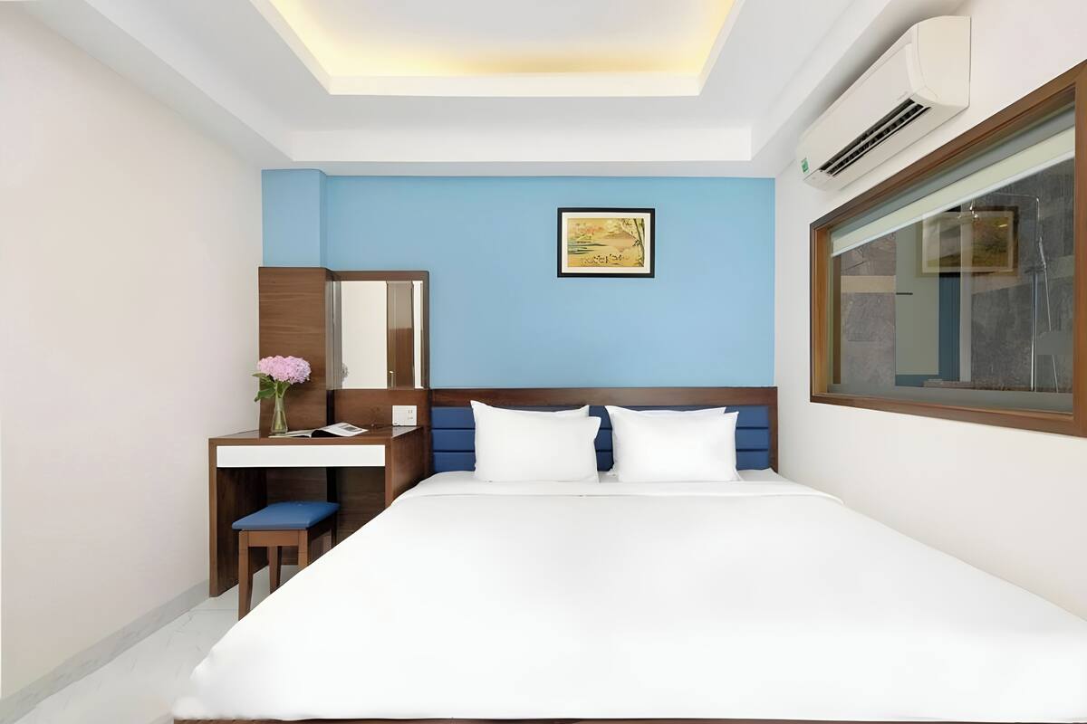 Habitación de hotel con cama, escritorio, silla, espejo, ventana y un cuadro en la pared.
