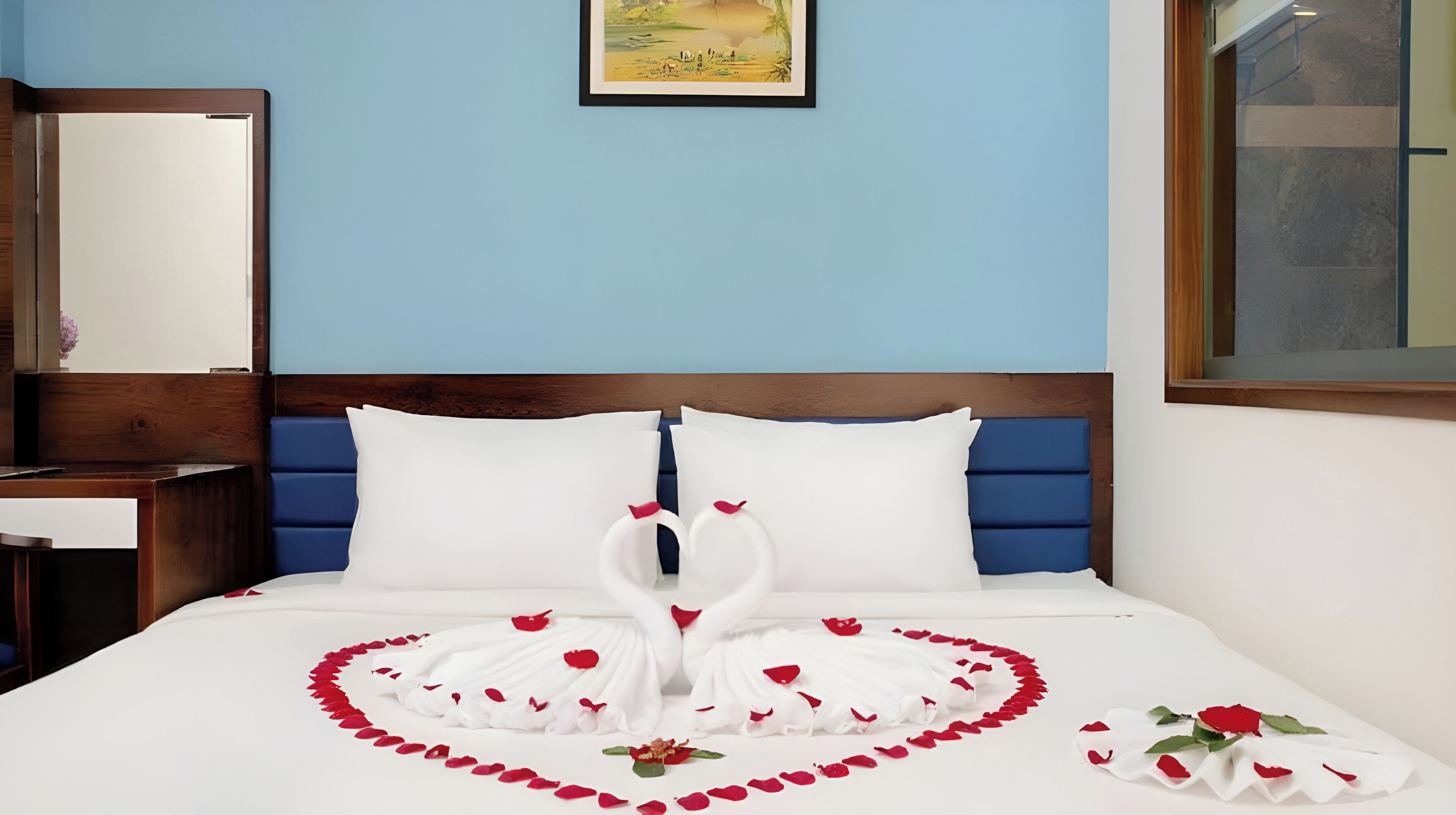 A neatly made bed with white pillows, a wooden headboard, and a heart-shaped decoration made of white fabric and red rose petals.