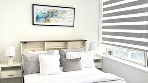 Une chambre à coucher bien rangée, avec un lit, des tables de chevet, une commode, un tableau encadré au mur et une fenêtre avec des stores.
