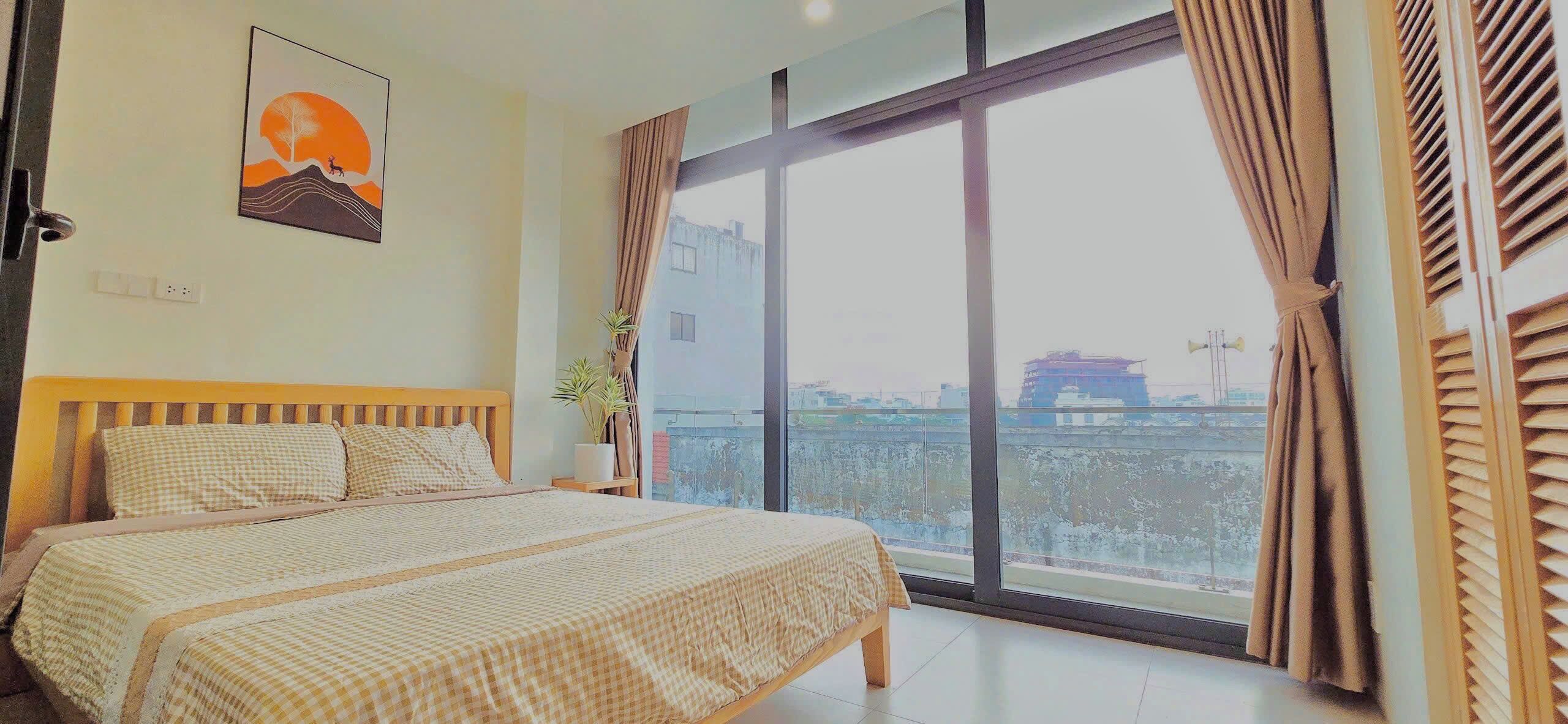 Un dormitorio con un ventanal grande, una cama con ropa de cama a cuadros, una mesita de noche, una planta en maceta y un cuadro colgado en la pared.