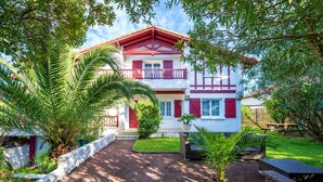 A two-story house with a red and white facade, a balcony, and a well-maintained garden with palm trees and a paved driveway. - Émilie's Guest Room - Anglet (Anglet)