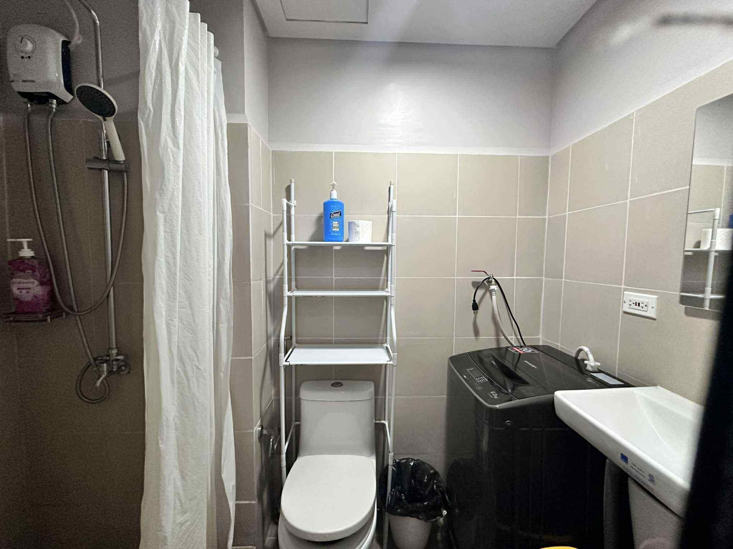 A compact bathroom with a toilet, a shower, and a sink. There are shelves above the toilet and a trash bin next to the sink.