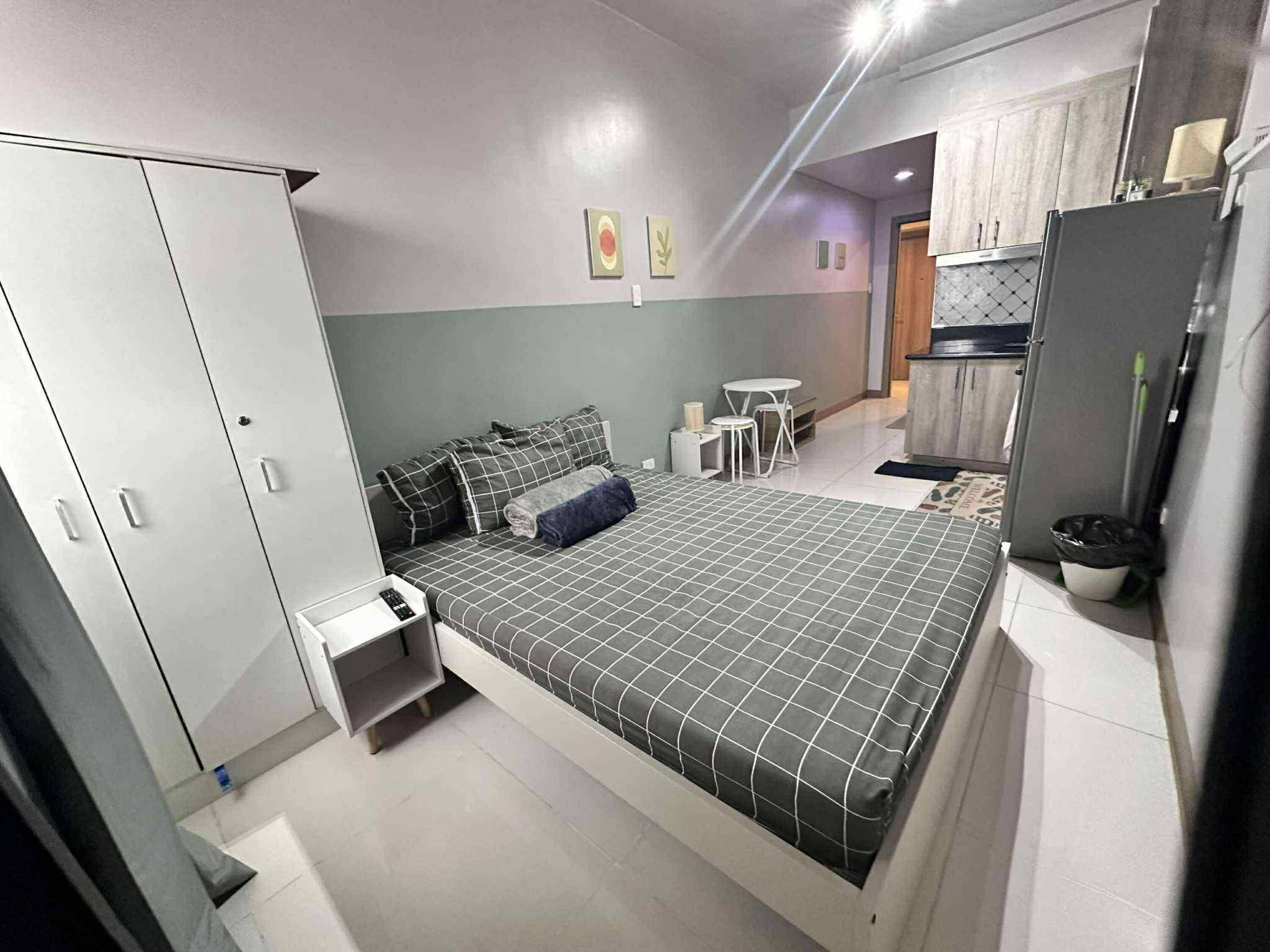 Quarto moderno com cama, guarda-roupa, mesa de cabeceira e pequena área de cozinha.