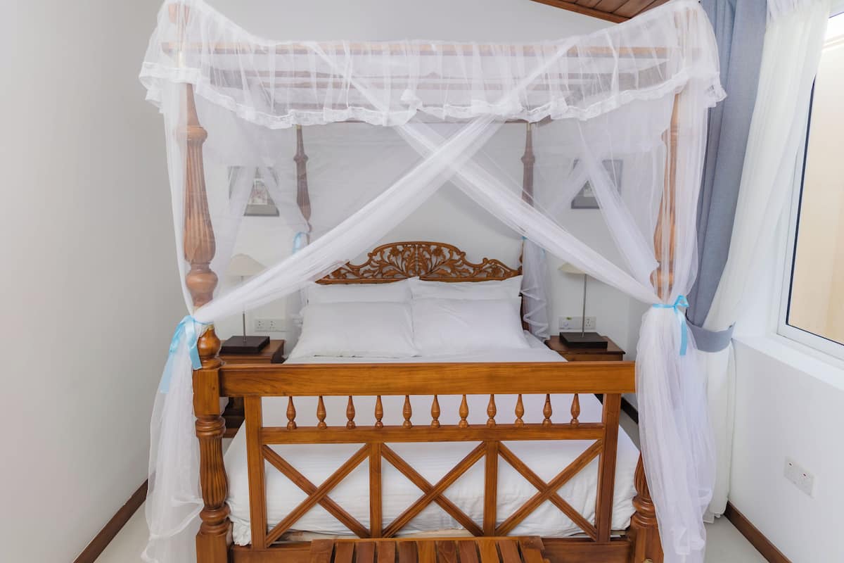A four-poster bed with a wooden frame and white bedding, a bedside table with a lamp, and a window with curtains.
