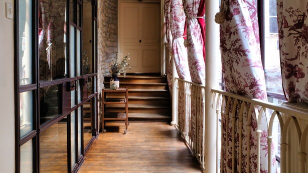Una scala in legno con una porta bianca in cima, un tavolino con un vaso di fiori e una ringhiera con tende floreali.