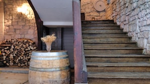 Una scala rustica con gradini in legno e un muro in pietra, che conduce a una stanza con una botte di legno e un vaso di fiori secchi.