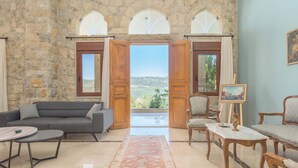 A spacious living area with a stone wall, a grey sofa, wooden chairs, a marble coffee table, and a view through open doors to a balcony. - Beit Salima 3BR House with Pool (Salima)
