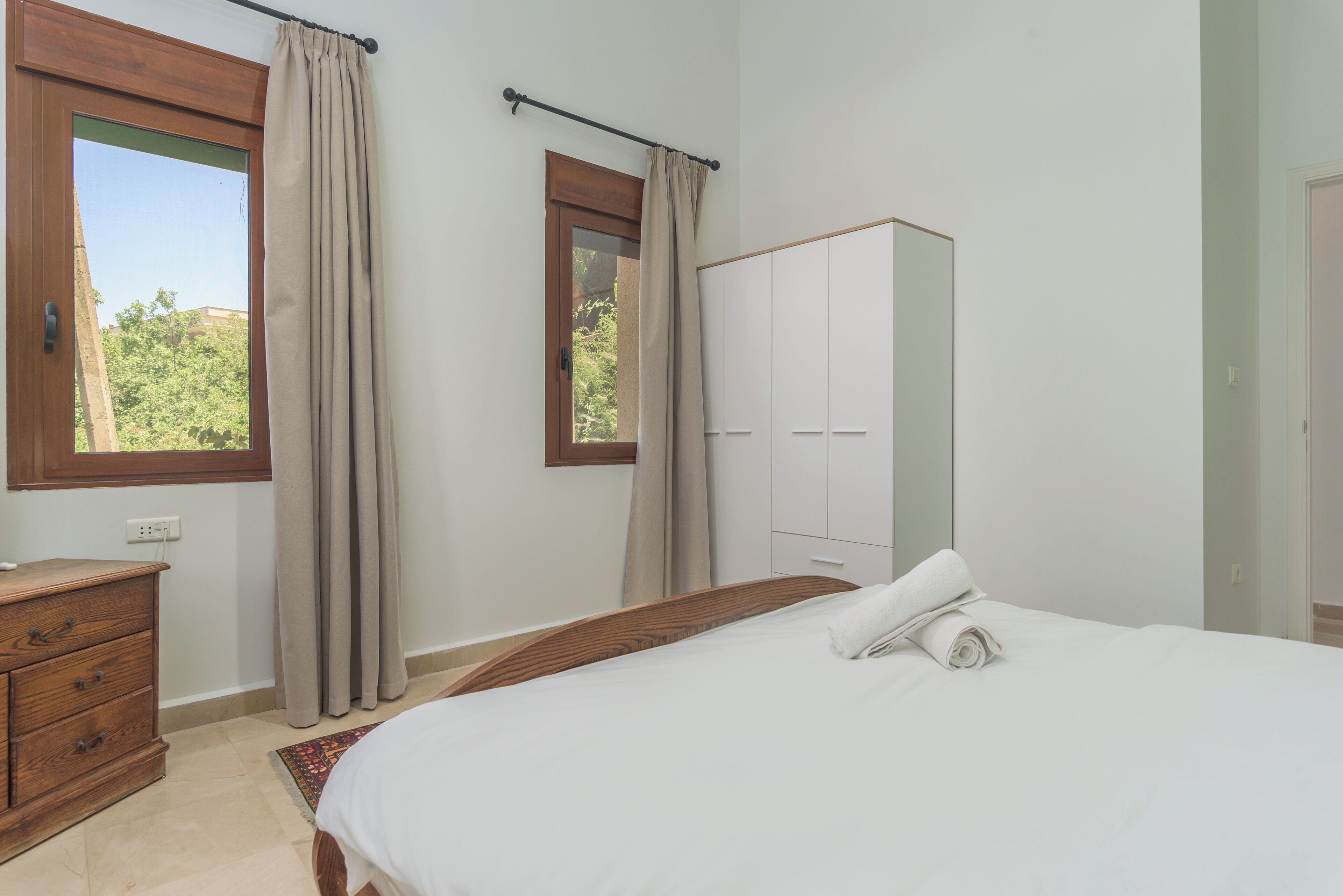 A bedroom with a bed, a wooden dresser, a wardrobe, and a window with curtains.