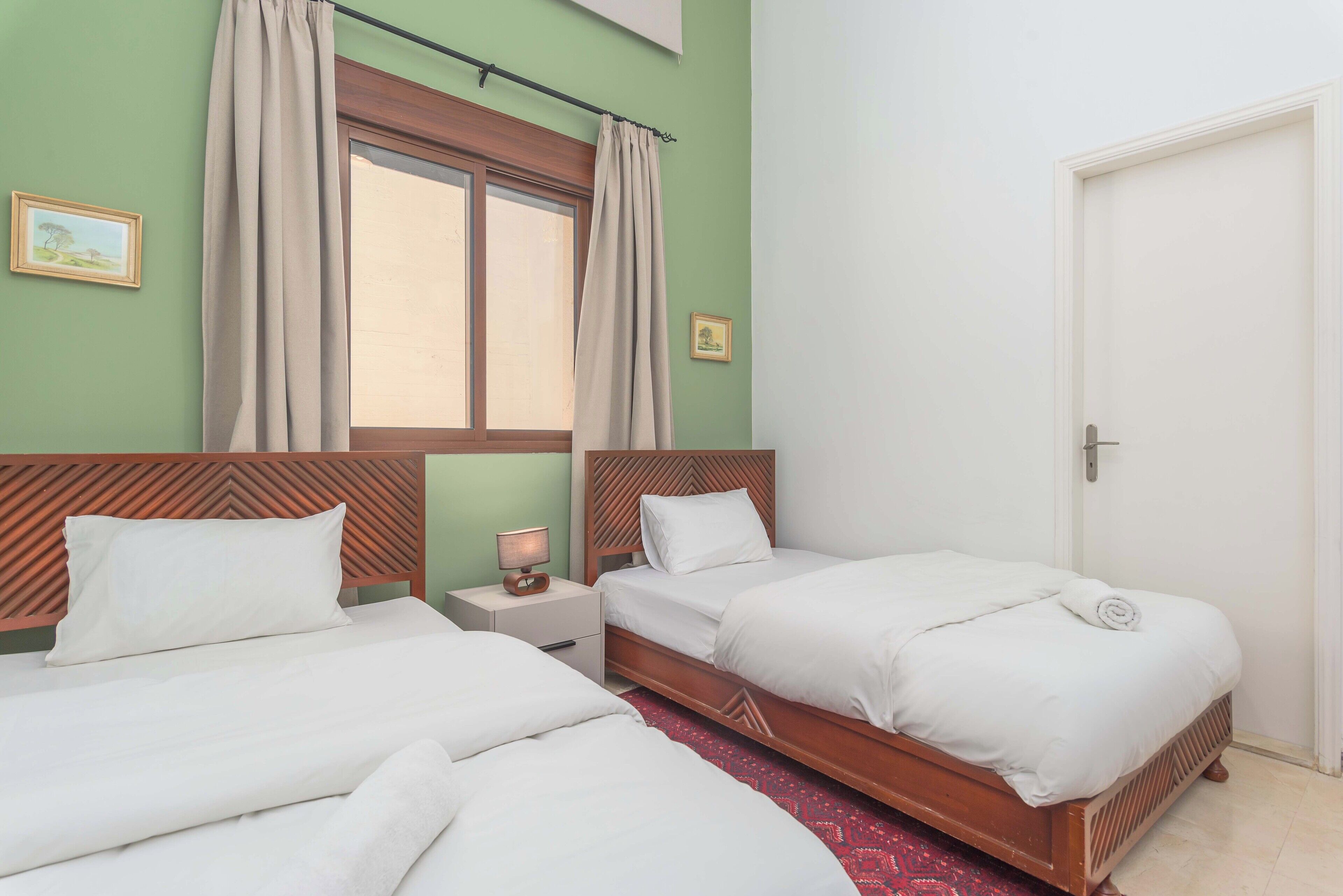 A hotel room with two beds, a window with curtains, a wooden headboard, a small bedside table with a lamp, and a door.