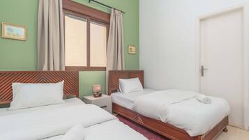 A hotel room with two beds, a window with curtains, a wooden headboard, a small bedside table with a lamp, and a door.