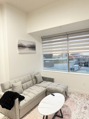A modern living room with a sofa, a coffee table, and a large window with blinds.