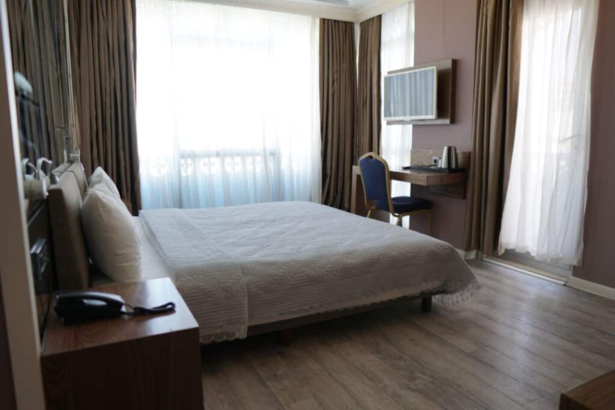 Een hotelkamer met een groot bed, een bureau met telefoon, een stoel, een televisie aan de muur en gordijnen voor de ramen.